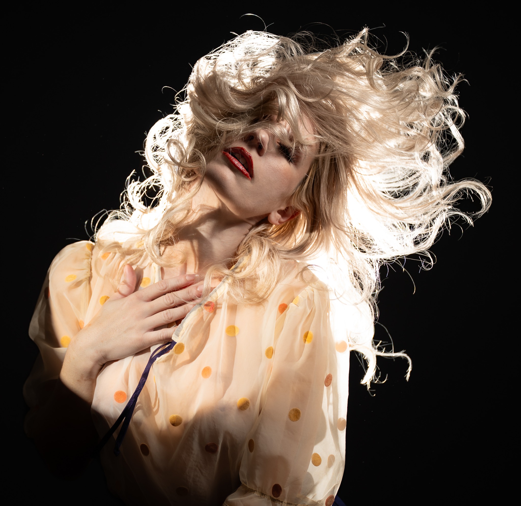 Dramatically backlit blonde curls in motion against black, with red lipstick and a cream polka-dot blouse